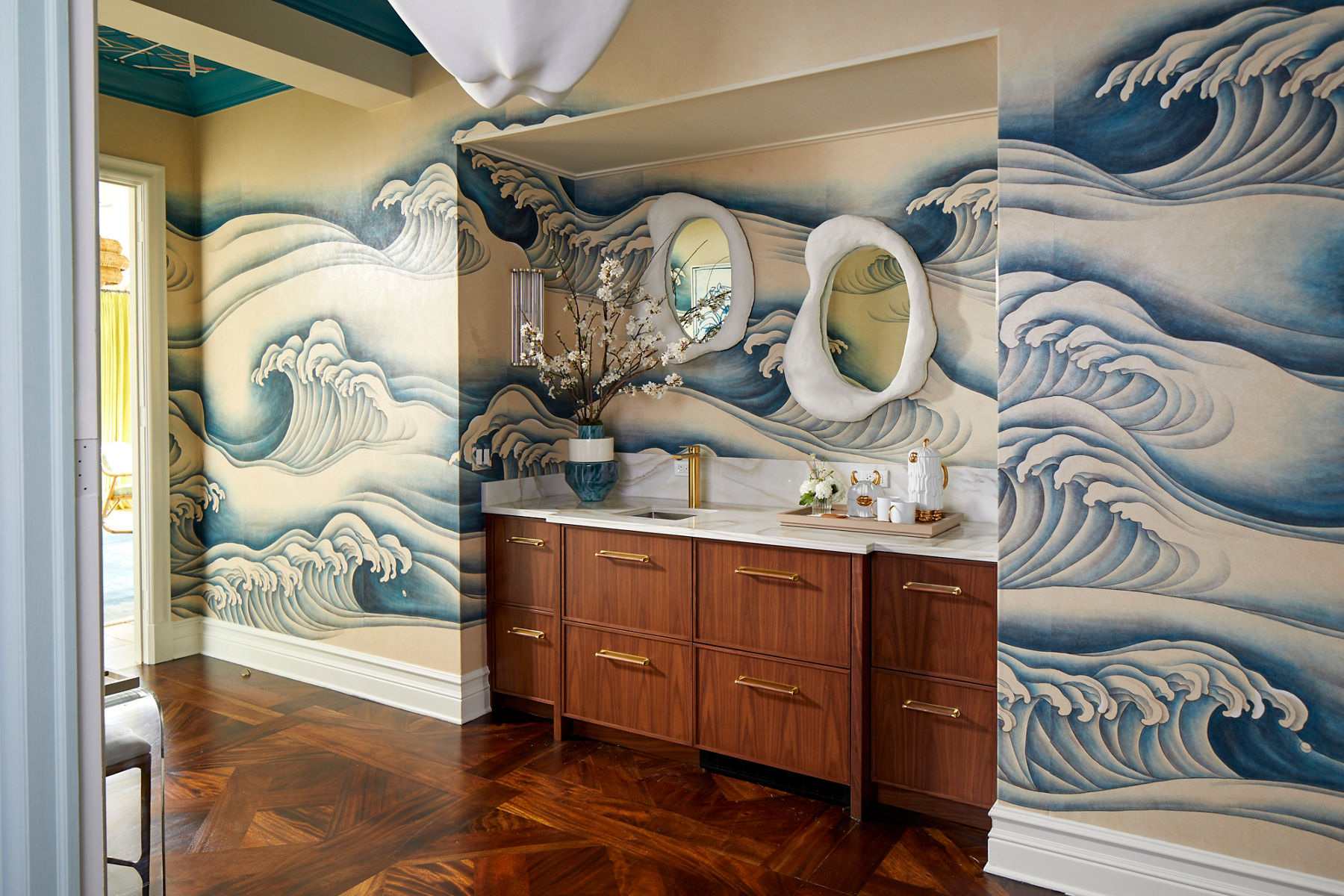 Wide image of a bedroom wall with large illustrated waves painted on the walls of the room. A wooden vanity and marble counter top are set into a small enclave along the wall. The space has a sink, brass faucet and circle mirror.