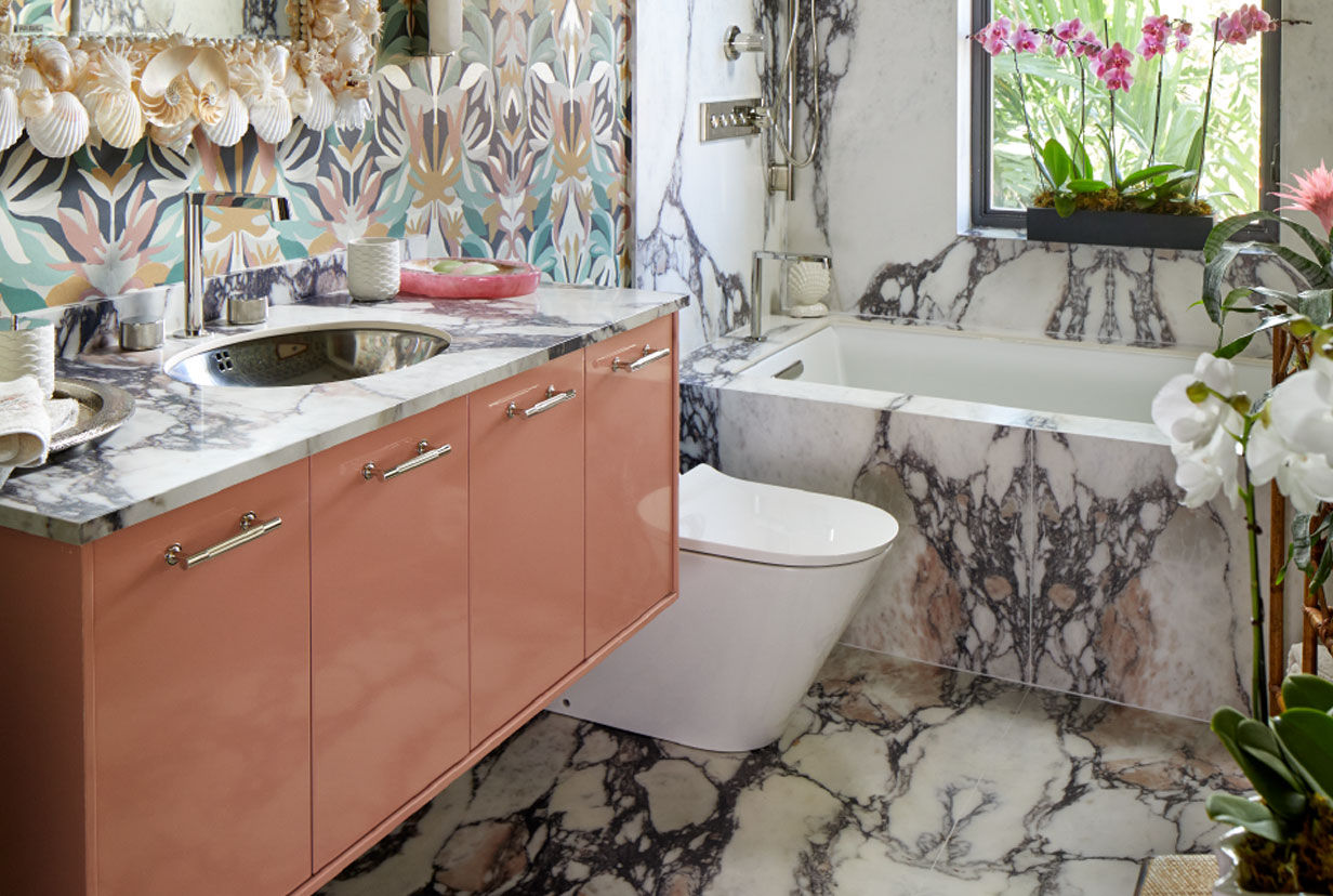 Image of an eclectic bathroom space with large, veined marble floor that wraps around the white bath. A matching marble slab vanity counter top features a stainless steel oval sink and Components Kohler faucet. Shells adorn the vanity mirror, which is set over a wildly colorful patterned wall.