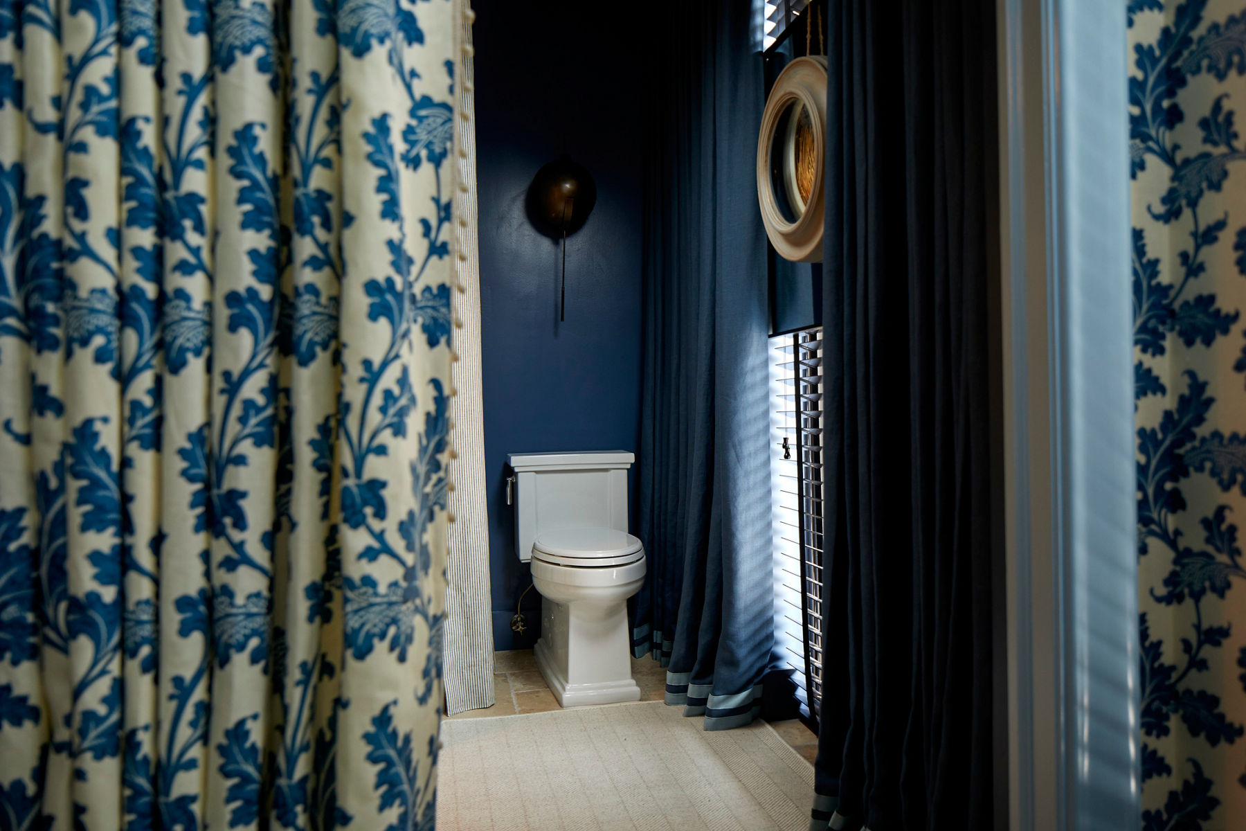 Image of a bathroom toileting space with focus on the white stately toilet. Navy blue walls have large patterned curtains of navy and white draped to the floor.