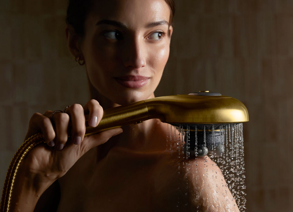 A close-up lifestyle image of a woman using a gold-toned body massager. The massager is placed on her right shoulder with water running.