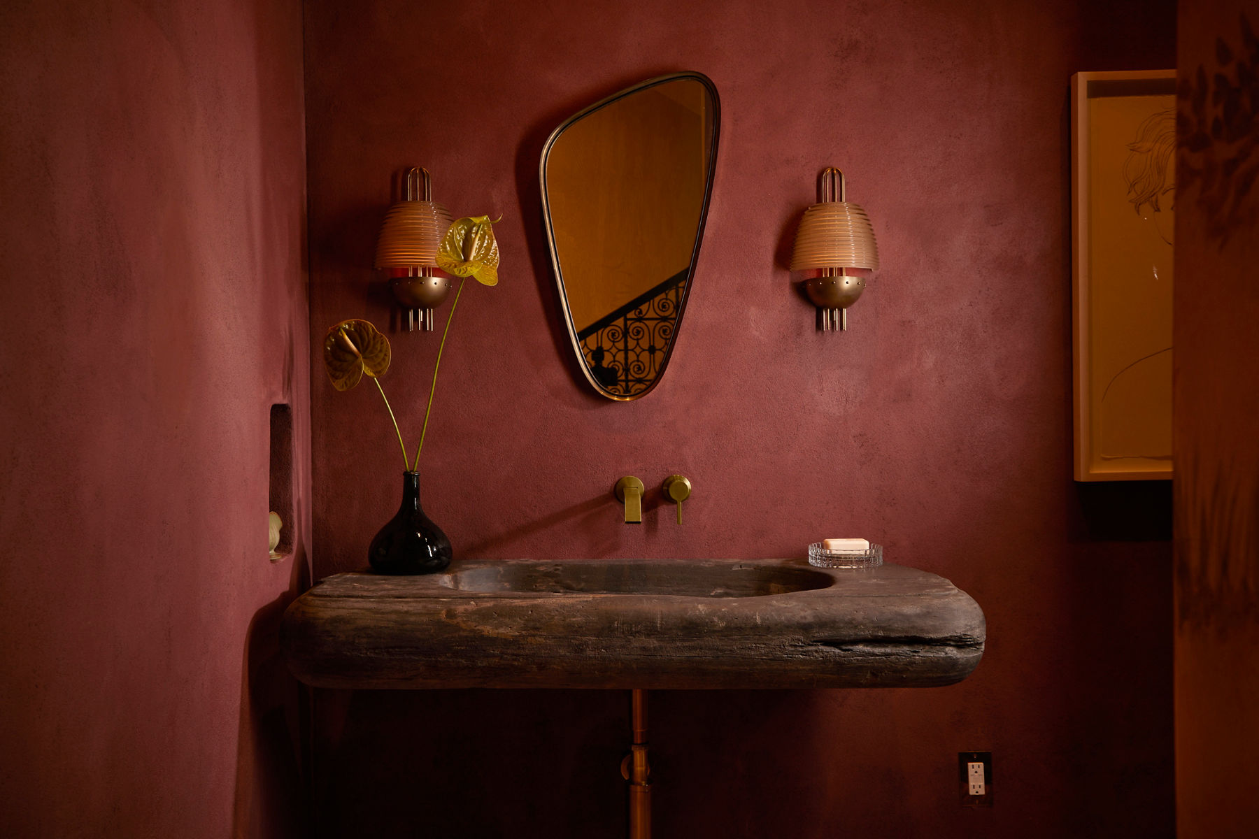 A sunlit maroon bathroom with orange sconces, an asymmetrical oval mirror, and a brown stone vanity.