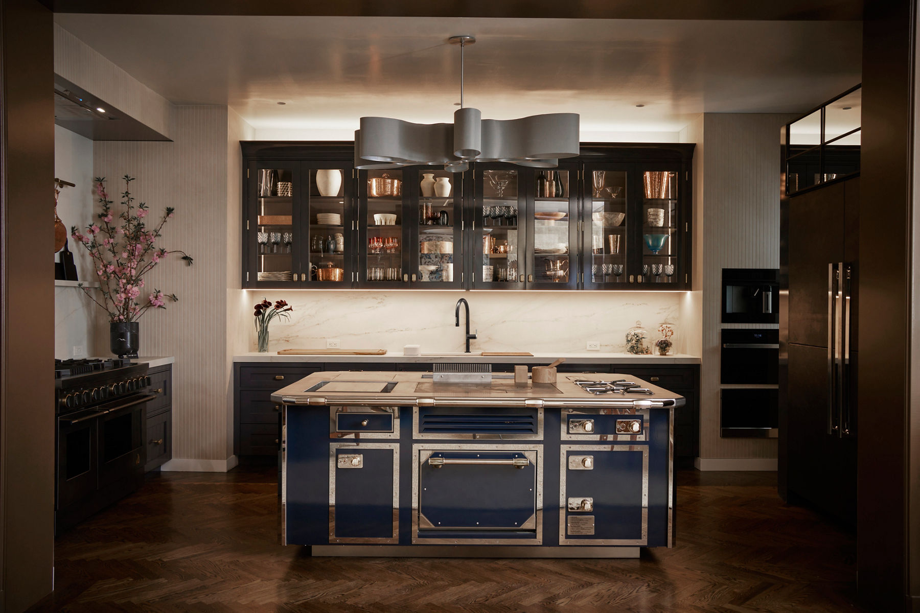 A dim lit kitchen with a treasure-chest inspired kitchen island and a kitchen counter and sink and faucet behind it. Theres glass shelving hanging from above.
