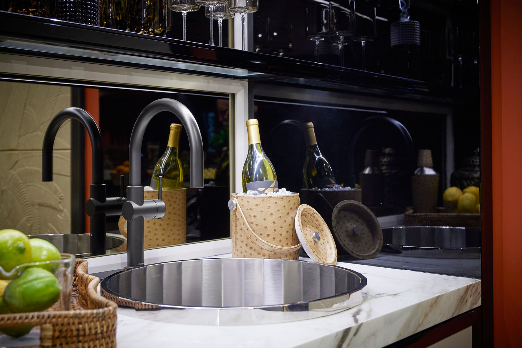 A close up shot of a bar sink designed in panneled silver with a black faucet. It's attached to a white marble countertop surrounded by a bottle of wine and bar accessories.