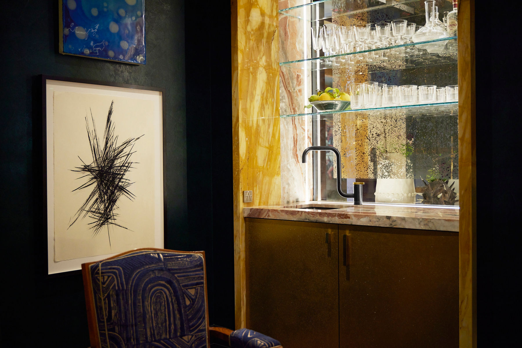 A moody bar space designed with large glass and mirrored marble cabinets with glasswear and decor on it. Two large paintings are on the lefthand side with an accent chair below it.