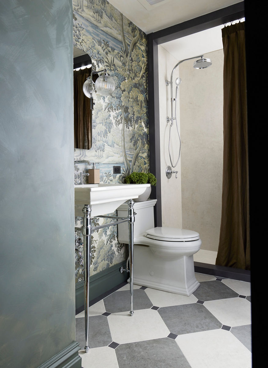 A calming bathroom space designed with sage green, floral wallpaper, a pale grey toilet, gray and white tiled floors, and a standing silver and white sink. There's a stand in shower in the background with beige walls and a dark brown shower curtain.
