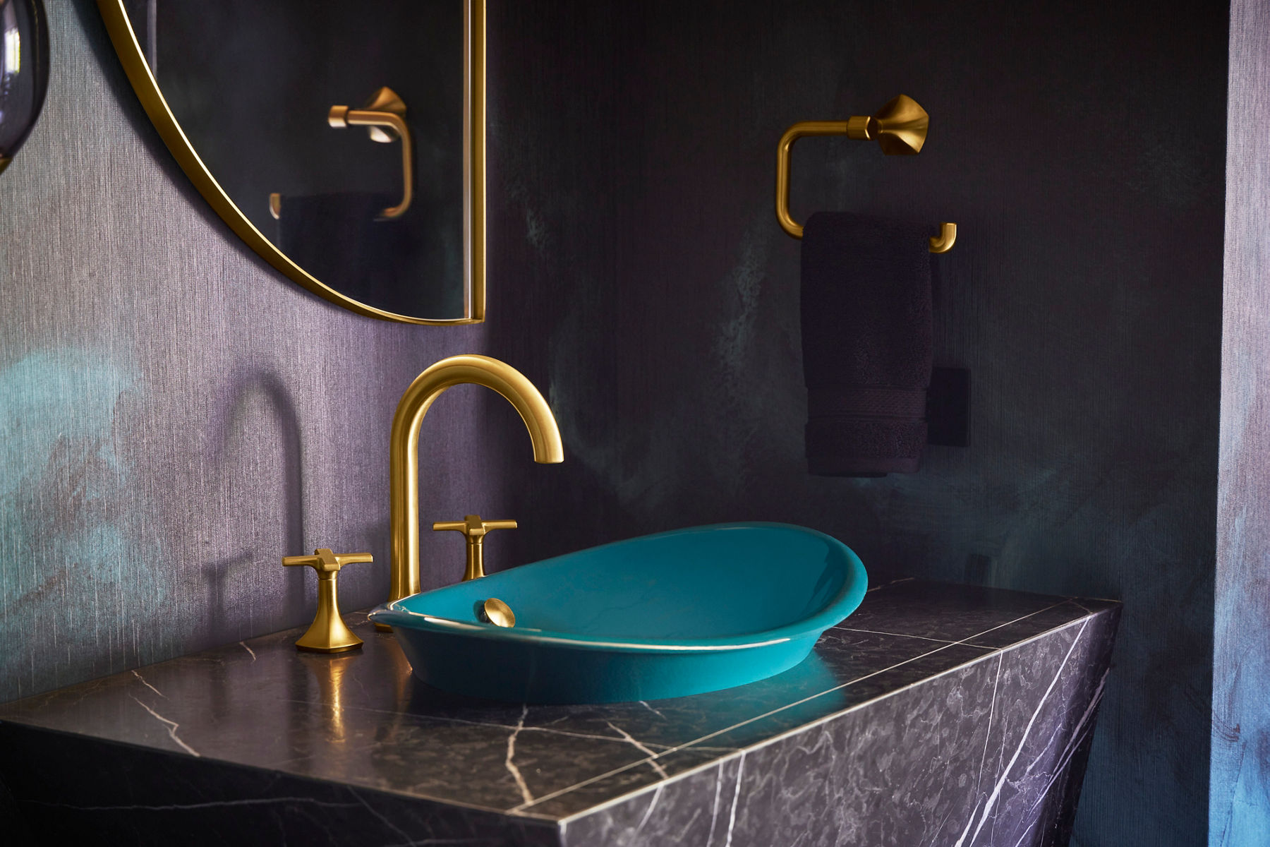 A teal bathroom in a moody and dramatically designed bathroom space. The floor is tiled and the wallpaper is an emerald green smoke design. There's a glass chandelier above and two large paintings against the back wall.