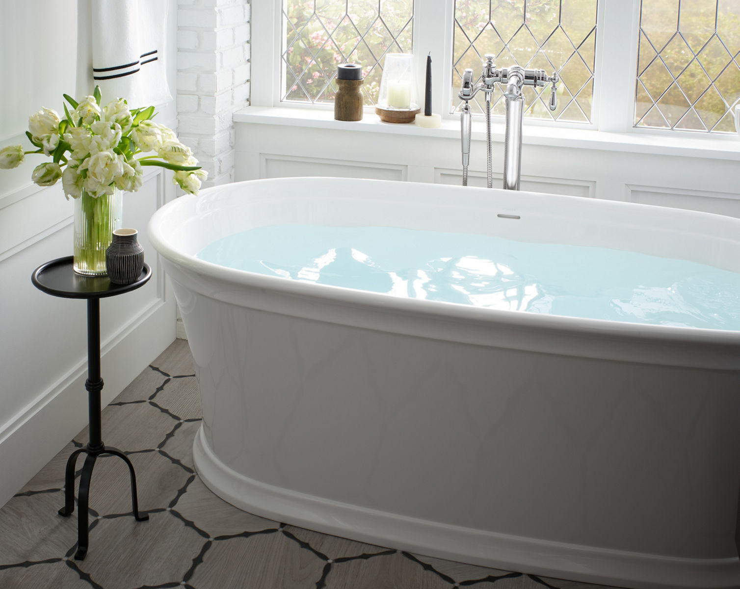 A vase of white flowers sits on a candlestick table beside a full white KOHLER freestanding bath with floor-mounted bathfiller and handshower.