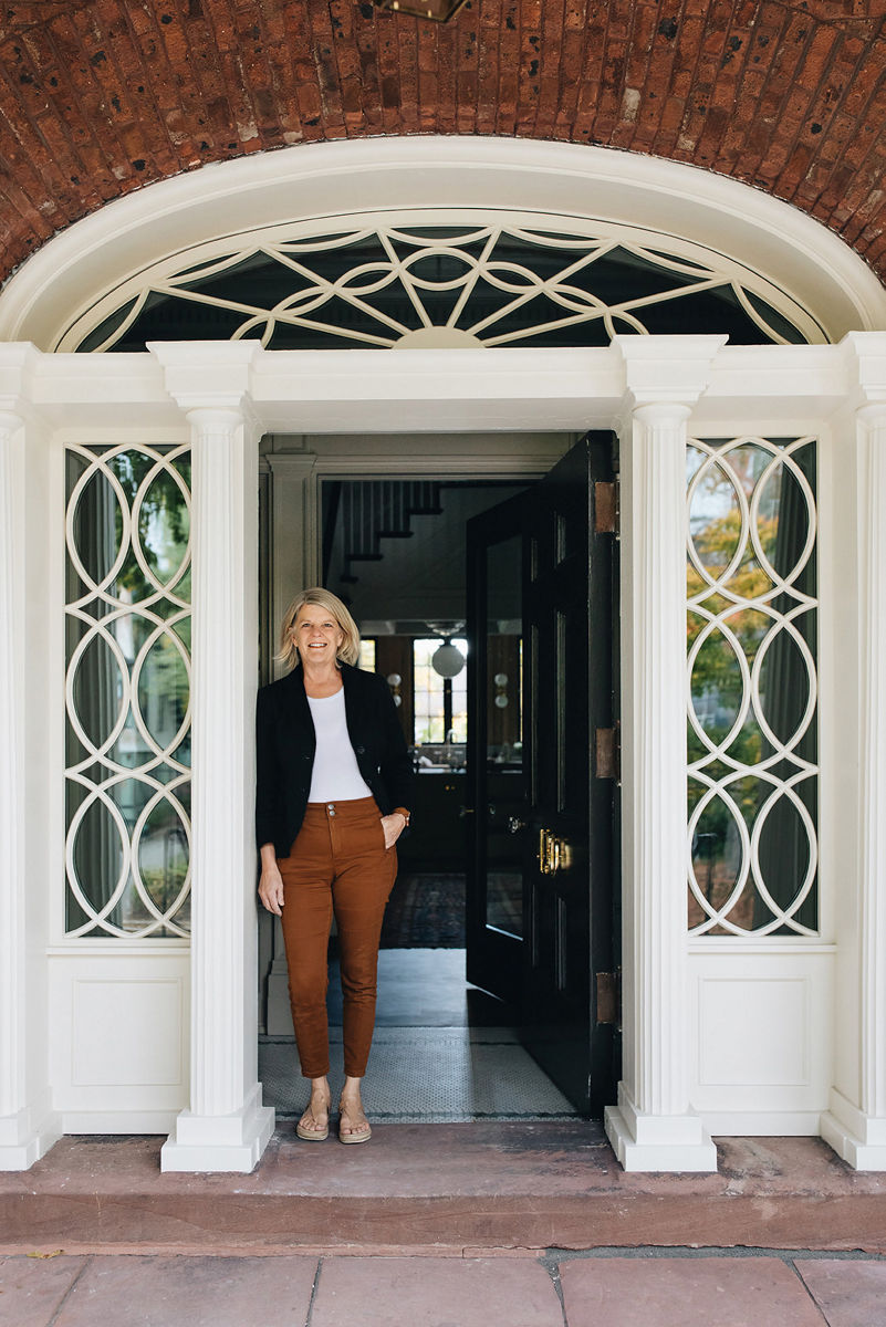 La designer Jean Stoffer, vêtue d’un pantalon marron, d’une chemise blanche et d’un blazer bleu foncé, s’appuie sur la porte ouverte de la maison qu’elle a conçue.