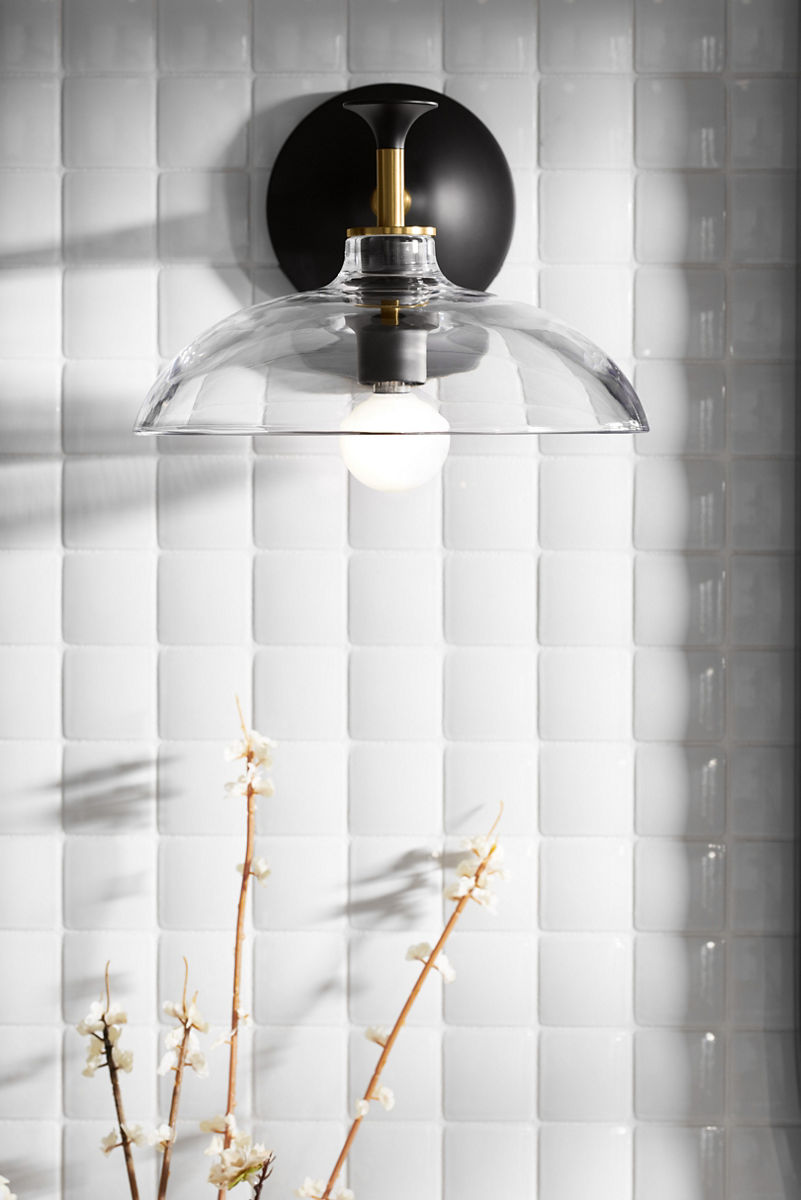 A KOHLER Tone sconce with glass shade on a white tiled wall.