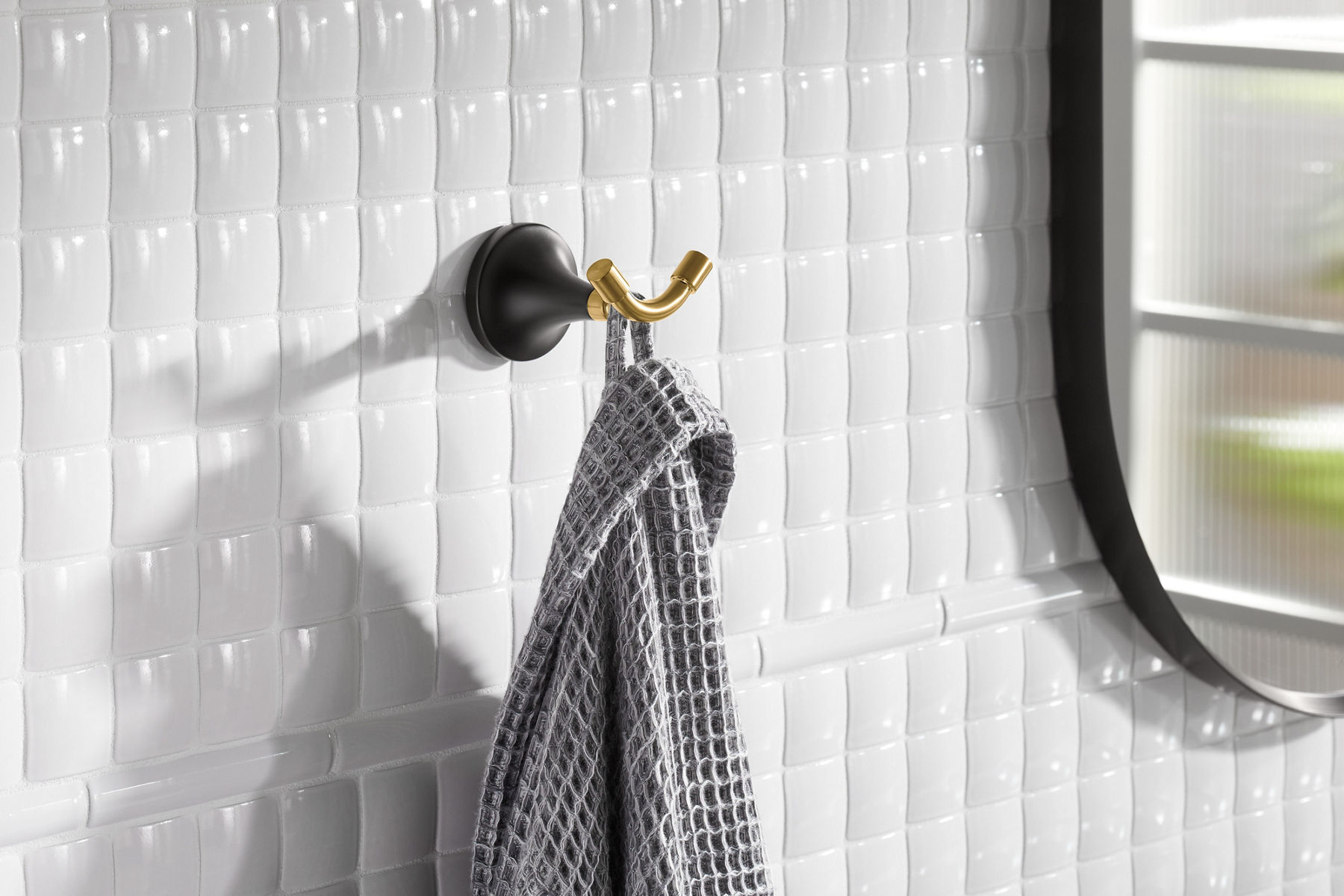 A gray waffle texture robe hangs on a KOHLER Tone robe hook mounted on a white tile wall.