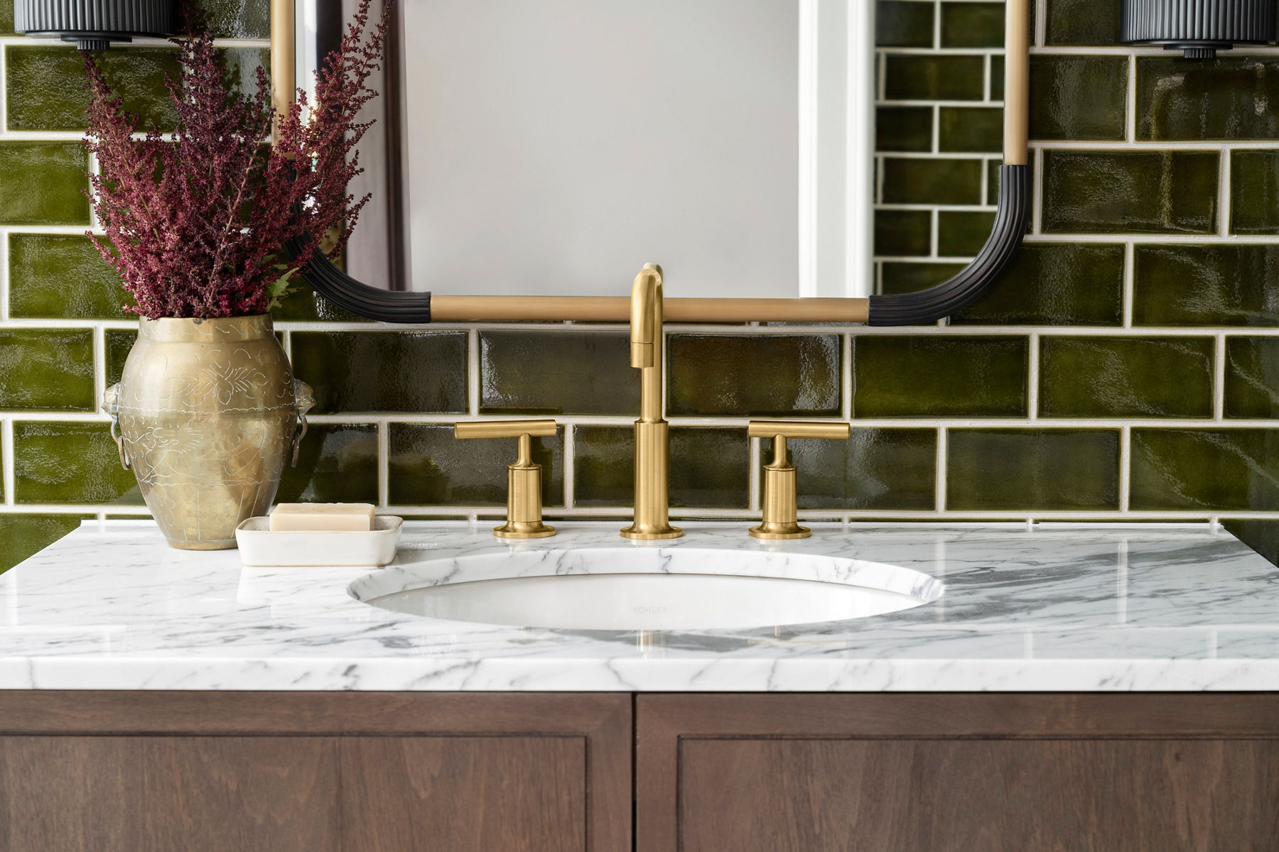 Brushed brass bathroom faucet with dual handles, mounted on a white marble countertop, set against a glossy green subway tile backsplash.