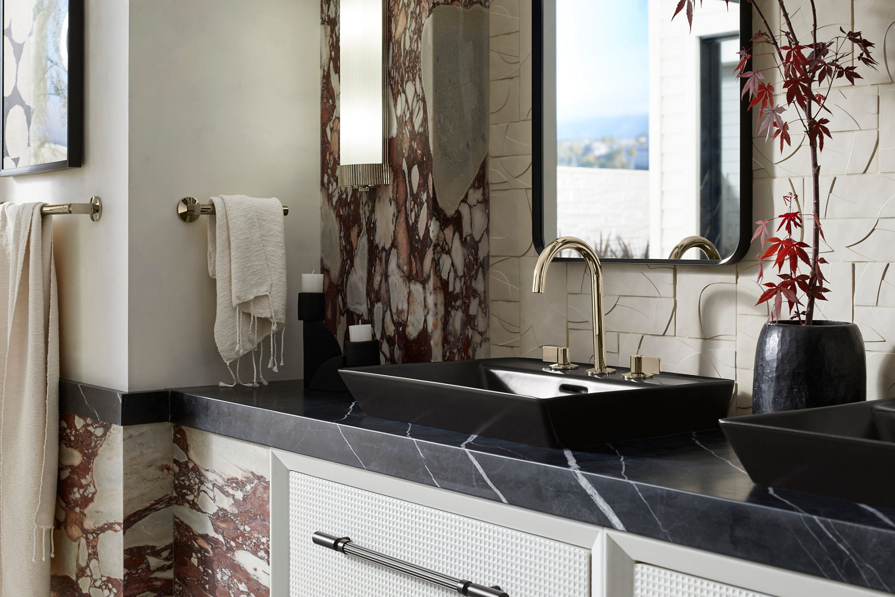 Luxurious bathroom vanity with a black rectangular vessel sink, brushed gold faucet, and a dark marble countertop, set against a textured wall with artistic tile and stone accents.