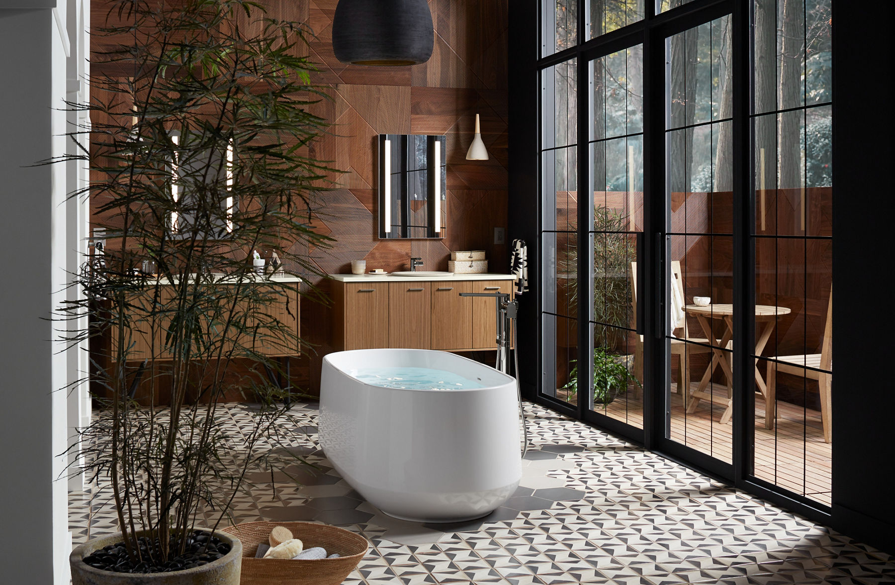 Une baignoire autoportante blanche sur un plancher en carrelage géométrique dans une salle de bain remplie de lumière avec de grandes portes vitrées à droite et deux espaces de meuble-lavabo en arrière-plan.