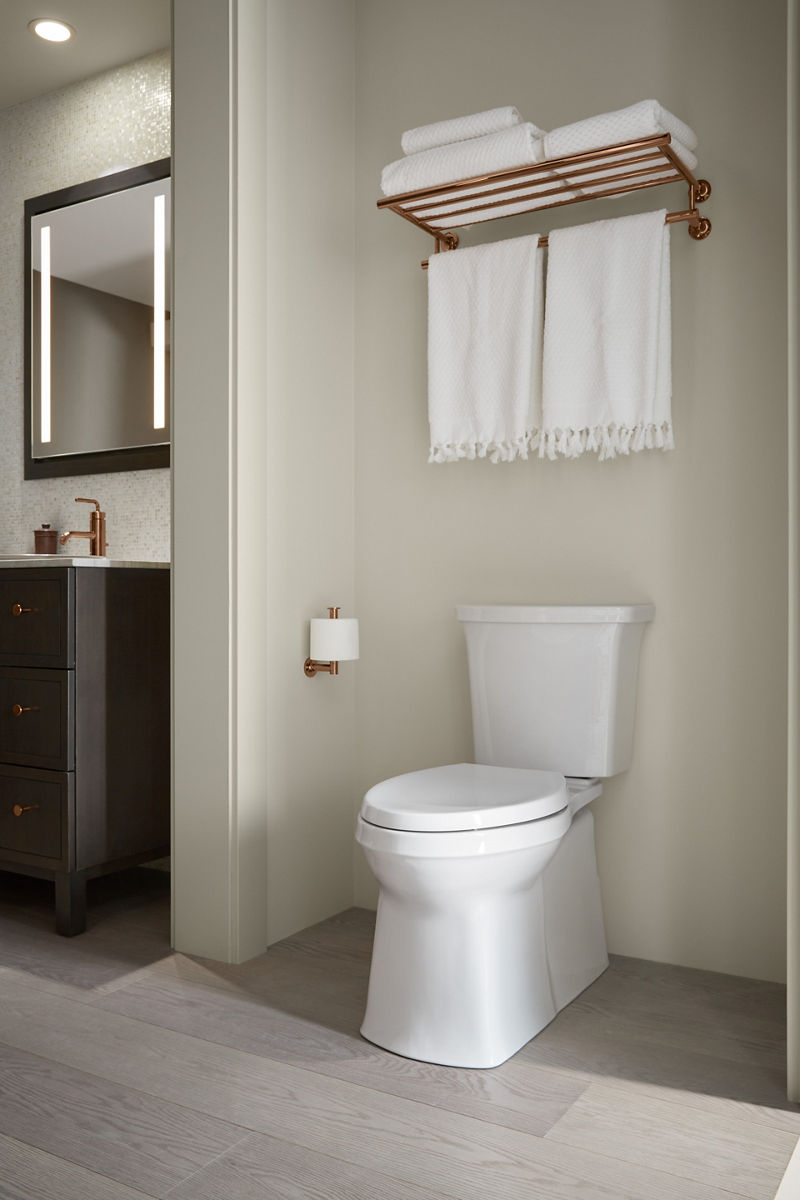 A white KOHLER Corbelle fully-skirted elongated toilet in a white bathroom with gray floors.