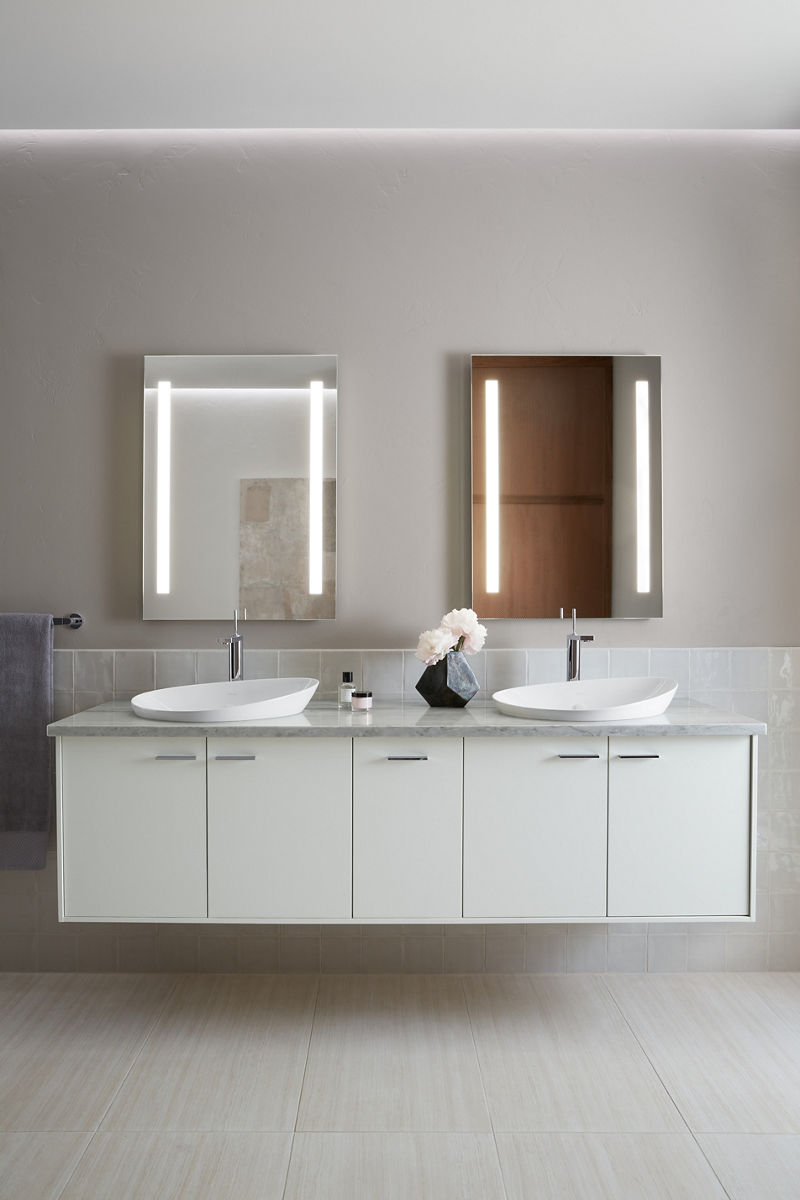 A lighted mirror with the two lights on the side of the mirror illuminated, sitting above a white vanity with stone counter and white sink.