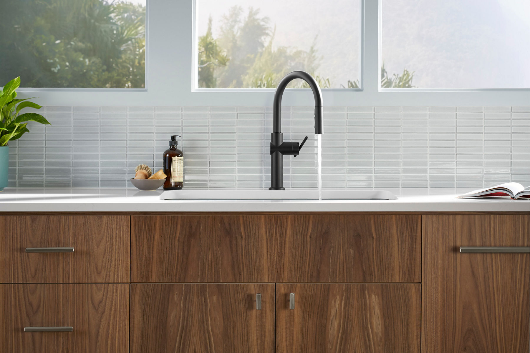 Matte black kitchen faucet with a curved spout and single lever handle, running water into a sink, set against a light horizontal tile backsplash.