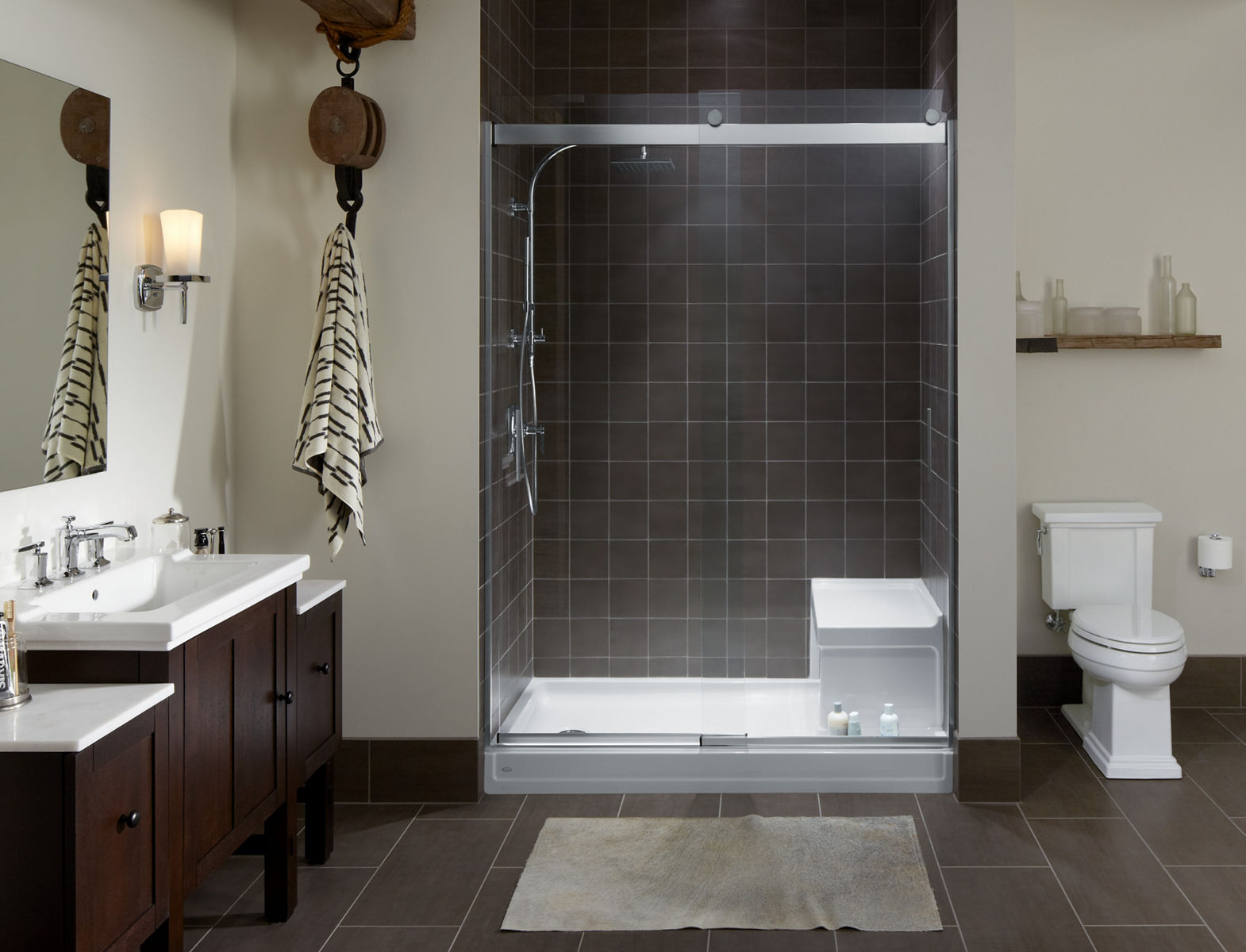 Une salle de bain avec une douche carrée grise avec une base de douche KOHLER Tresham avec siège intégré, des portes de douche sans cadre Levity, une colonne de douche et une douchette.