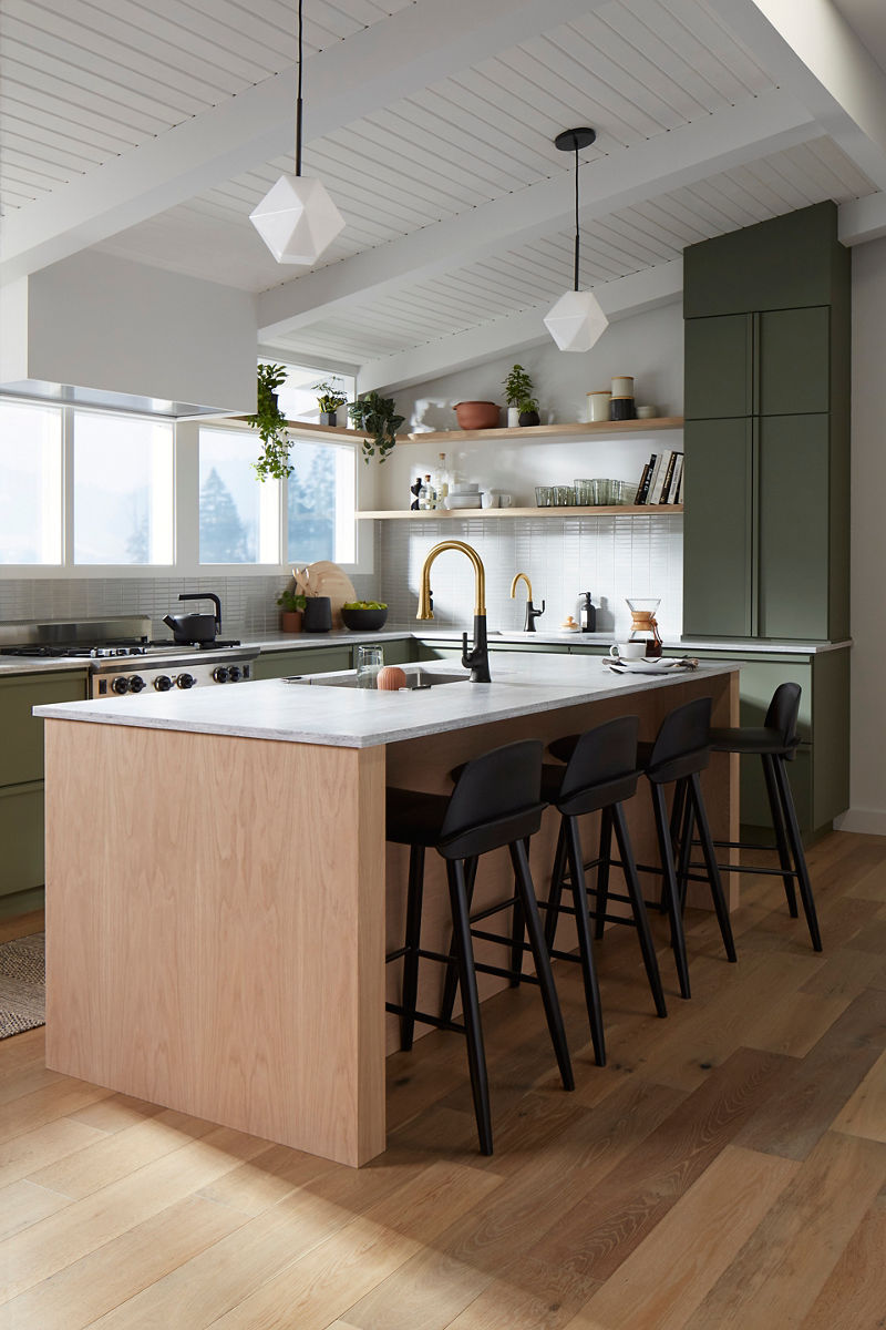 Wide image of a sunny kitchen with earthy green cabinet colors and natural wood. The large island features a centered sink with two-tone Kohler faucet in the center, across from a wall of windows and open storage shelves.