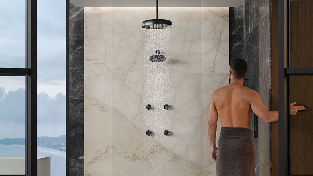 Banner image featuring a luxurious marble shower with a matte black rain showerhead and body jets in use, with water cascading down. A man, seen from behind, is entering the shower, and the space is framed by large glass windows offering a view of the cloudy sky.