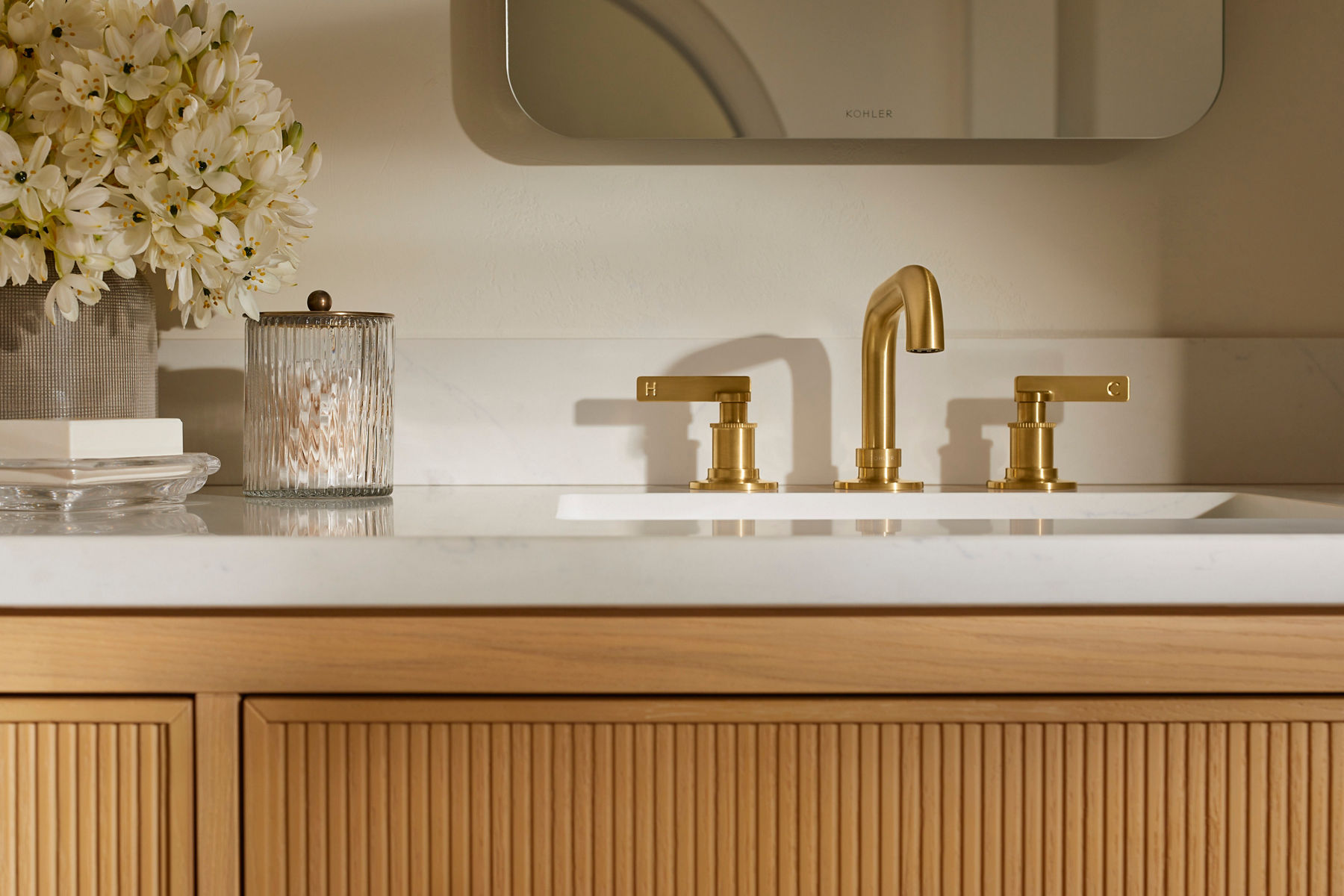 Un dessus de meuble-lavabo pour salle de bain avec un robinet de salle de bain Casita Kohler au fini doré et des poignées à levier, avec d’autres accessoires de meuble-lavabo en arrière-plan