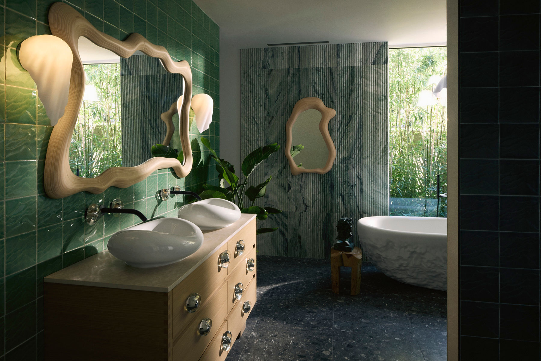 A modern bathroom showcasing the Kohler x Arsham Landshapes™ Collection. The image features a white sculptural sink on a wooden vanity, a uniquely shaped wooden mirror, and green tiled walls. In the background, a freestanding white bathtub is partially visible next to large windows that allow natural light to filter in.