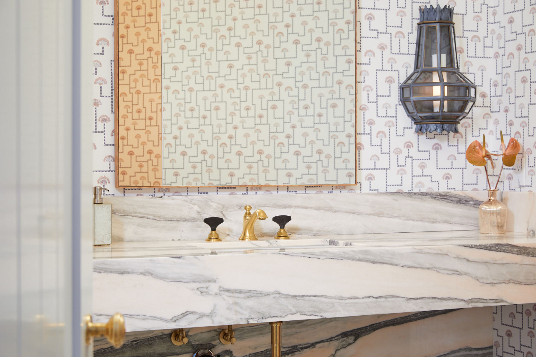 Gros plan sur le robinet de salle de bain de la salle d’eau Reese au fini bronze brillant avec poignées aux accents de pierre foncée. Le robinet se trouve au-dessus d’un lavabo carré et d’un comptoir en pierre de ton neutre. On peut voir un papier peint à imprimés autour du miroir.