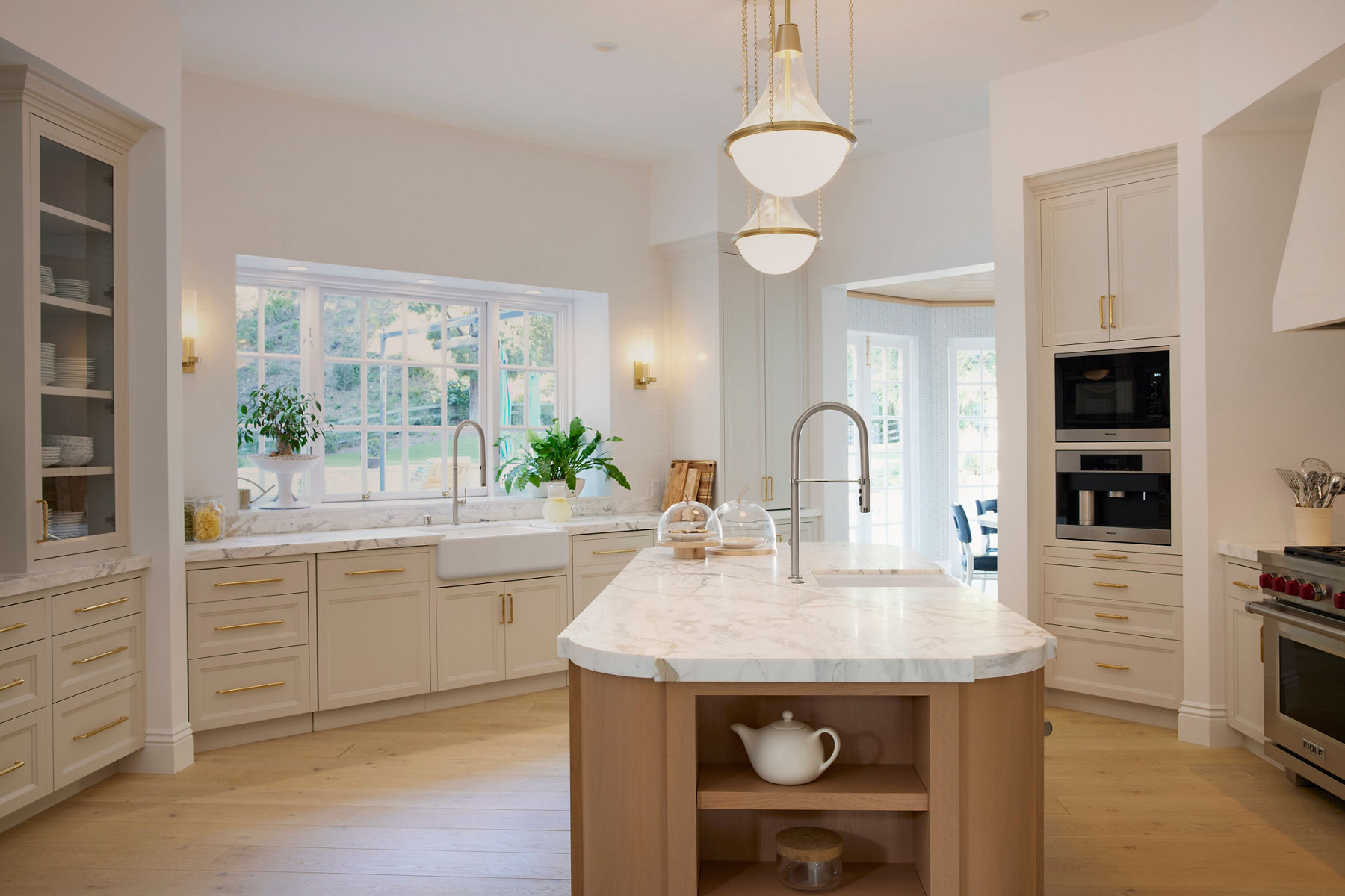 Wide angle of Reese's kitchen with two matching tall semi-professional faucets in Brushed Nickel finish. One faucet is above a sink at the kitchen island. The other is above a white farmhouse sink in front of the kitchen window. Cream cabinets and walls have brass pulls and accessories. 