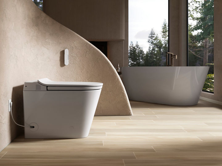 Stylish bathroom featuring a modern white smart toilet and a freestanding bathtub. The toilet is positioned near a curved, textured beige wall with a small control panel mounted beside it. The floor is light wood, and a large window offers a view of a forested landscape, adding natural light to the space.