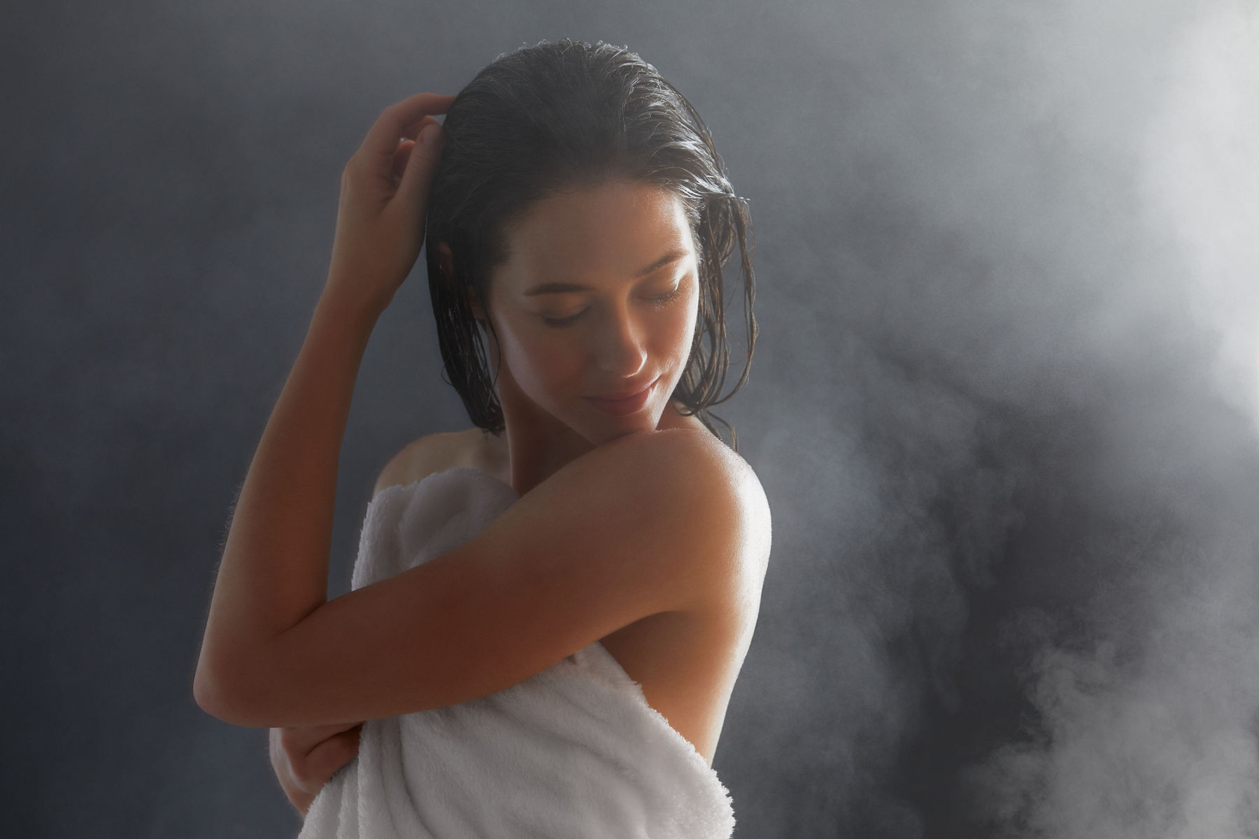 Gros plan d’une femme qui vient de sortir de la douche avec les cheveux mouillés et une serviette enroulée autour d’elle, alors qu’elle regarde en arrière la peau hydratée de son épaule et du haut de son dos.  