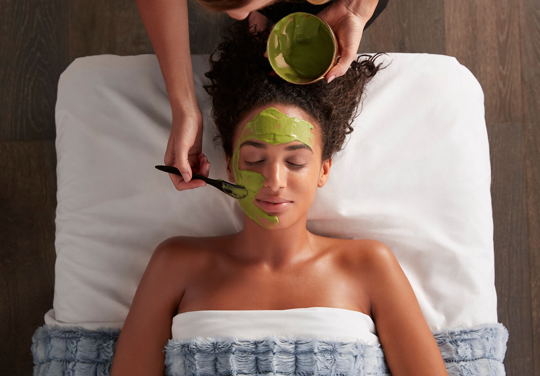 Overhead image of a woman laying face-up on a spa table with her eyes close, as she's having a mask facial treatment applied with a brush by a spa professional.