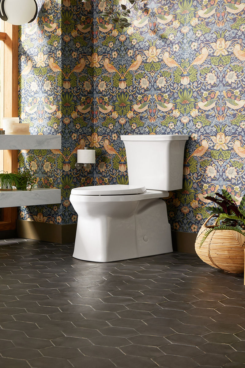 Modern white toilet in a bathroom with decorative floral wallpaper and dark hexagonal floor tiles.