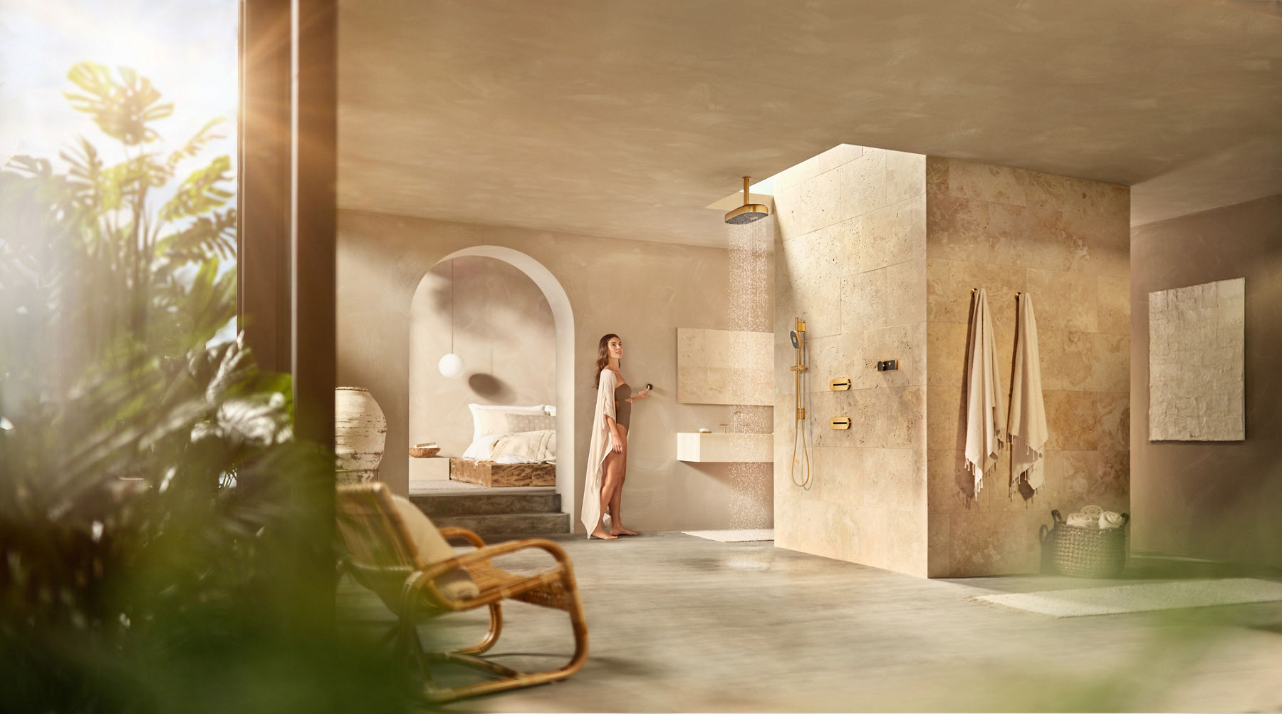 Une femme dans une grande salle de bain avec des carreaux de pierre du plancher au plafond se tient à distance pendant qu’elle actionne la pomme de douche à effet de pluie.