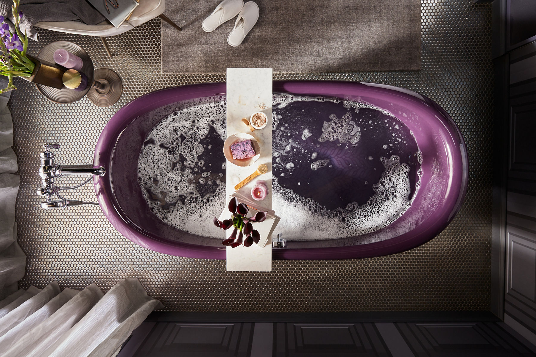 Overhead view of a KOHLER Artifacts freestanding bath with Black Plum exterior, bath tray holding flowers, a candle, soap, and bath salts with floor-mount bath filler trim with handshower.