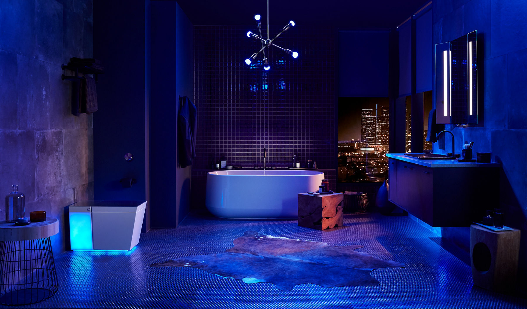 A large, darkened bathroom illuminated by a bluish, purple light with a Numi toilet on the left, a freestanding bath in the center background and a wall-mount vanity and internally lit mirror on the right.