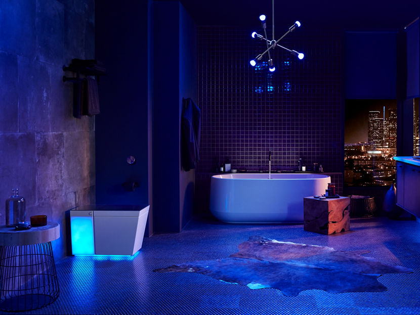 A large, darkened bathroom illuminated by a bluish, purple light with a Numi toilet on the left, a freestanding bath in the center background and a wall-mount vanity and internally lit mirror on the right.