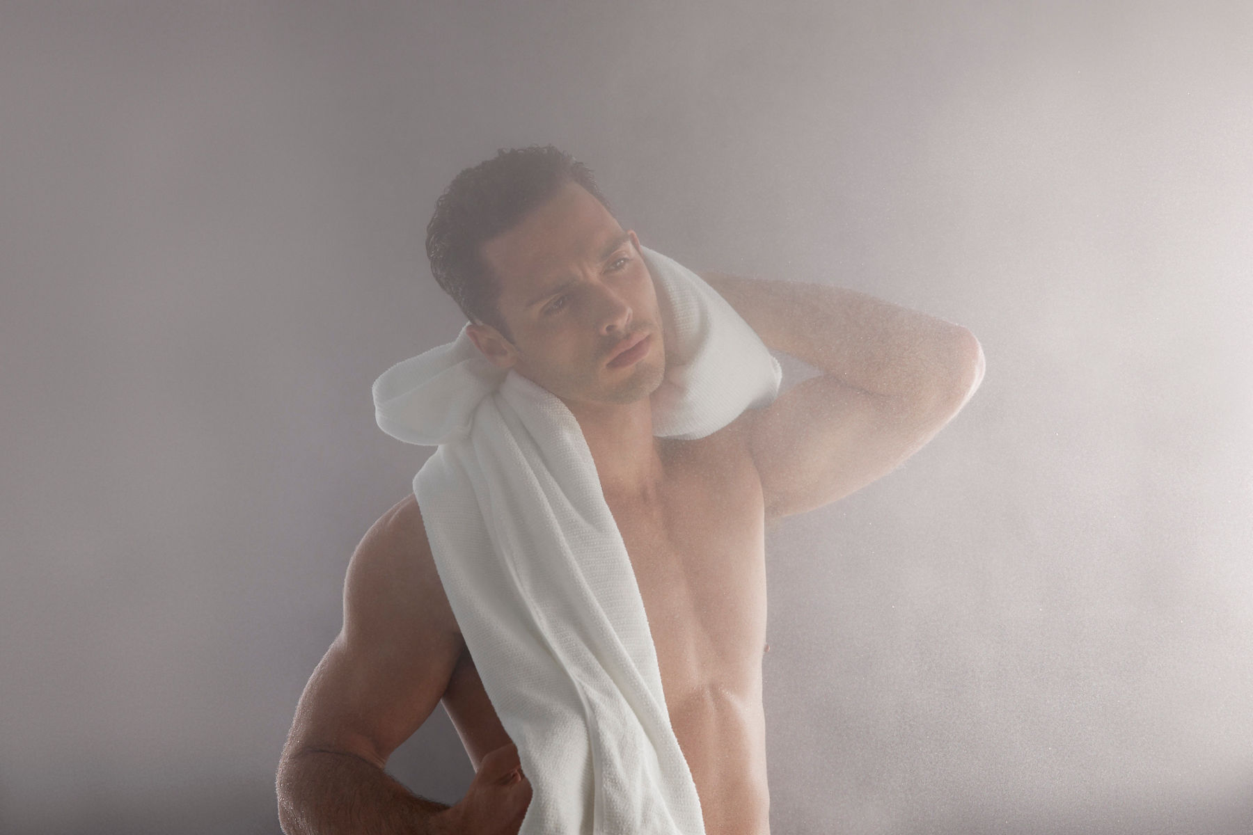 Portrait d’un homme dans une pièce embuée se séchant la nuque avec une serviette blanche drapée sur son épaule.