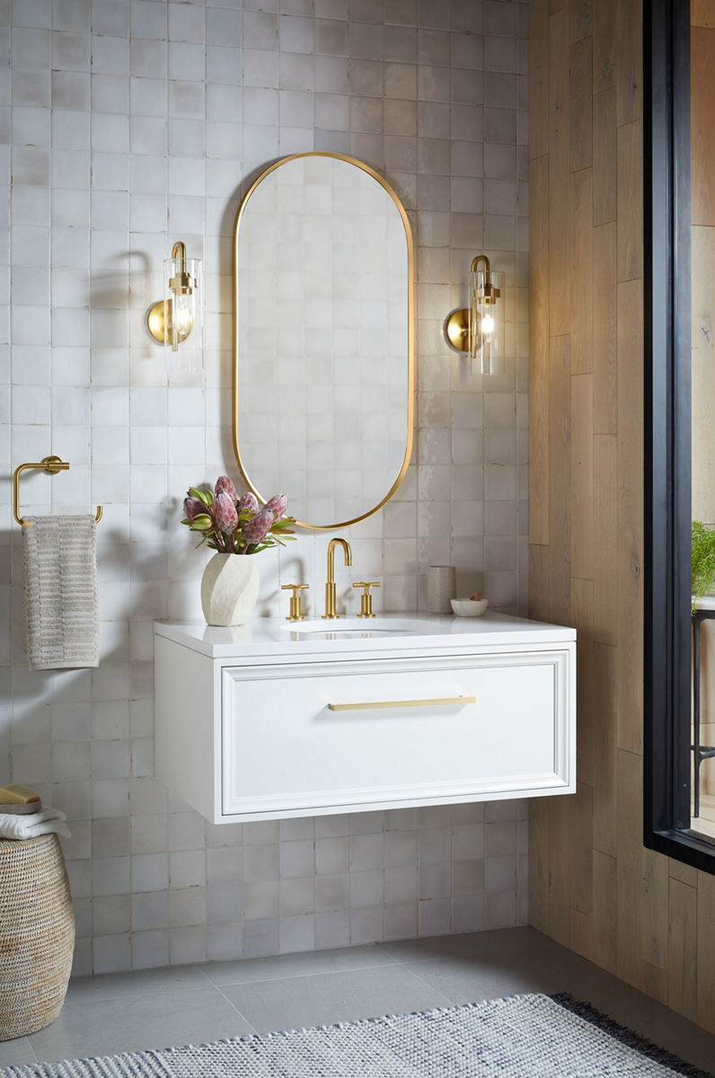 Un robinet de lavabo de salle de bain déployé Purist de KOHLER avec col de cygne et poignées en croix installé sur un dessus de meuble-lavabo blanc sous un miroir en forme de capsule.