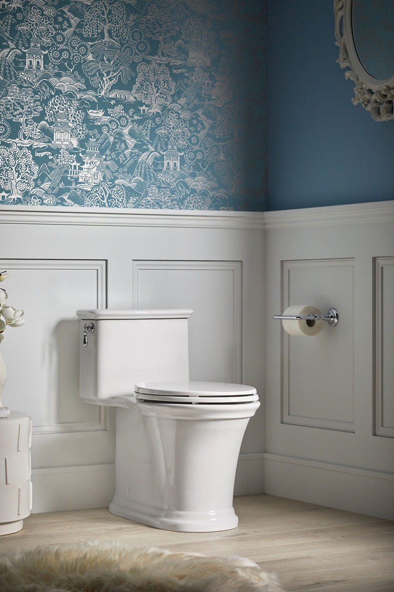 A serene bathroom with a white KOHLER one-piece toilet, wood floors, and silver and blue wallpaper above white wainscoting.