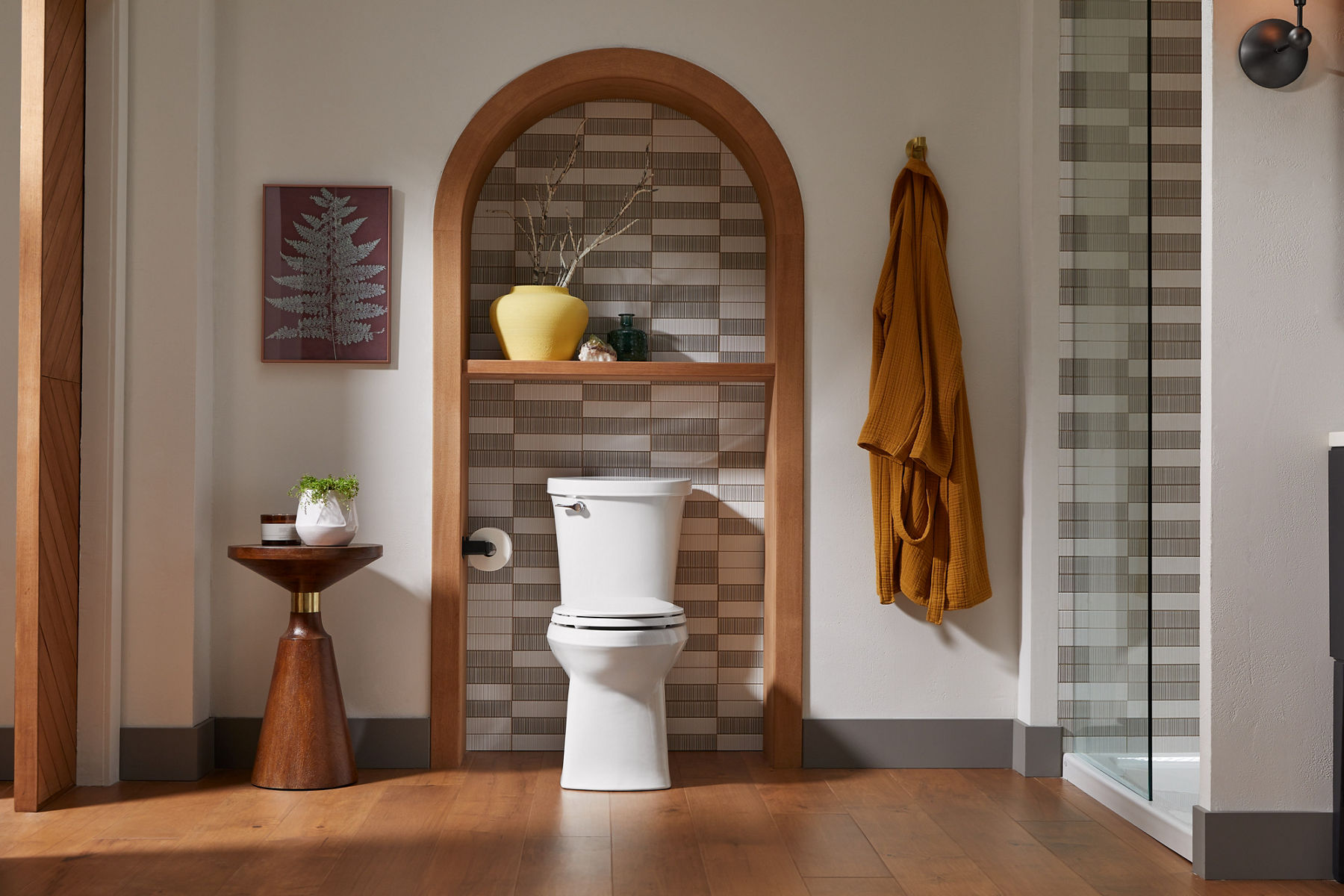 A bathroom with white KOHLER toilet installed in an arched white and gray tiled niche, accent shelving, wall art, and a robe hanging from a hook.