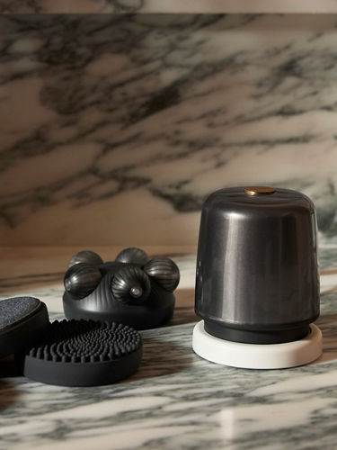 A portrait lifestyle image of bath products placed on a marble counter. The products include a black body massager and interchangeable attachments.