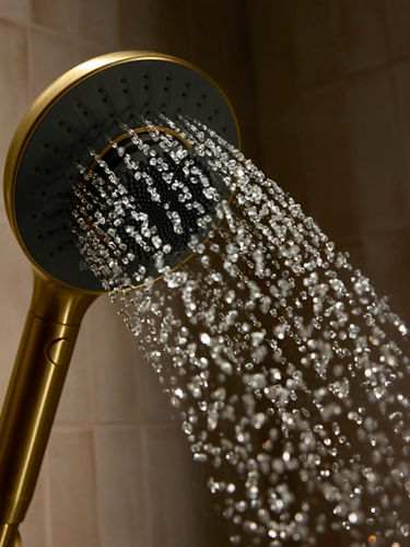 A close-up portrait product image of a gold and black running shower head attached to a beige tiled wall.