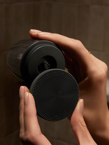 A close-up product shot of a compact black facial brush. The facial brush is opened up and being held by two hands.