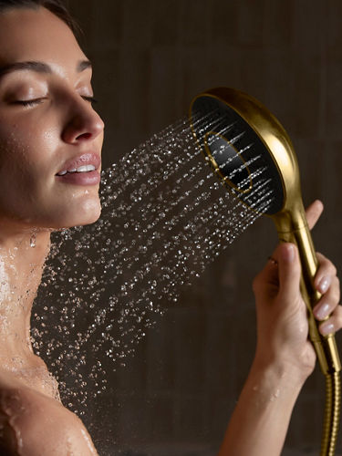 A close-up portrait shot of a woman using a gold-toned shower head. The shower head is placed on her neck with water running.