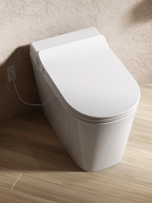 Close-up of a sleek, modern white smart toilet with a closed lid, positioned against a textured beige wall. The toilet is plugged into a nearby electrical outlet, and the floor is covered with light wood planks, complementing the minimalist design of the bathroom.