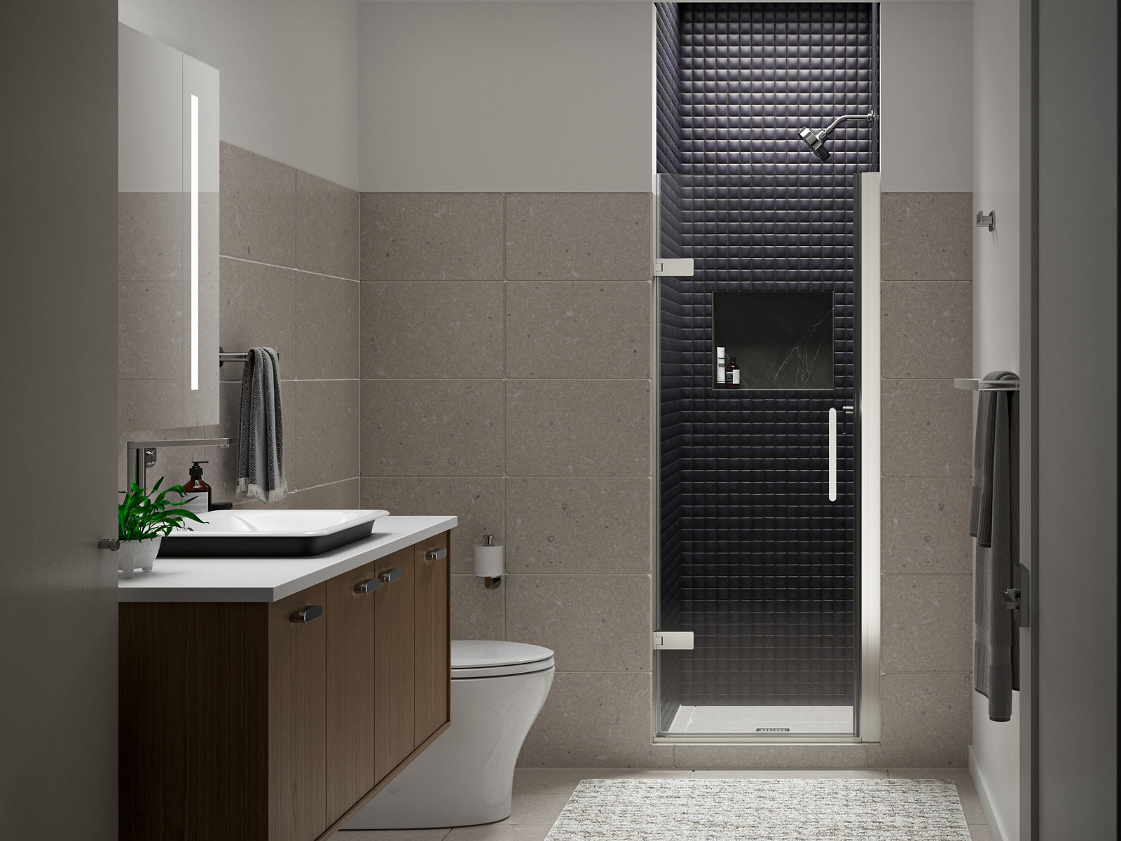 Douche carrelée gris foncé avec porte de douche pivotante en verre KOHLER à l’extrémité d’une salle de bain aux murs blancs et beiges et meuble-lavabo mural brun.