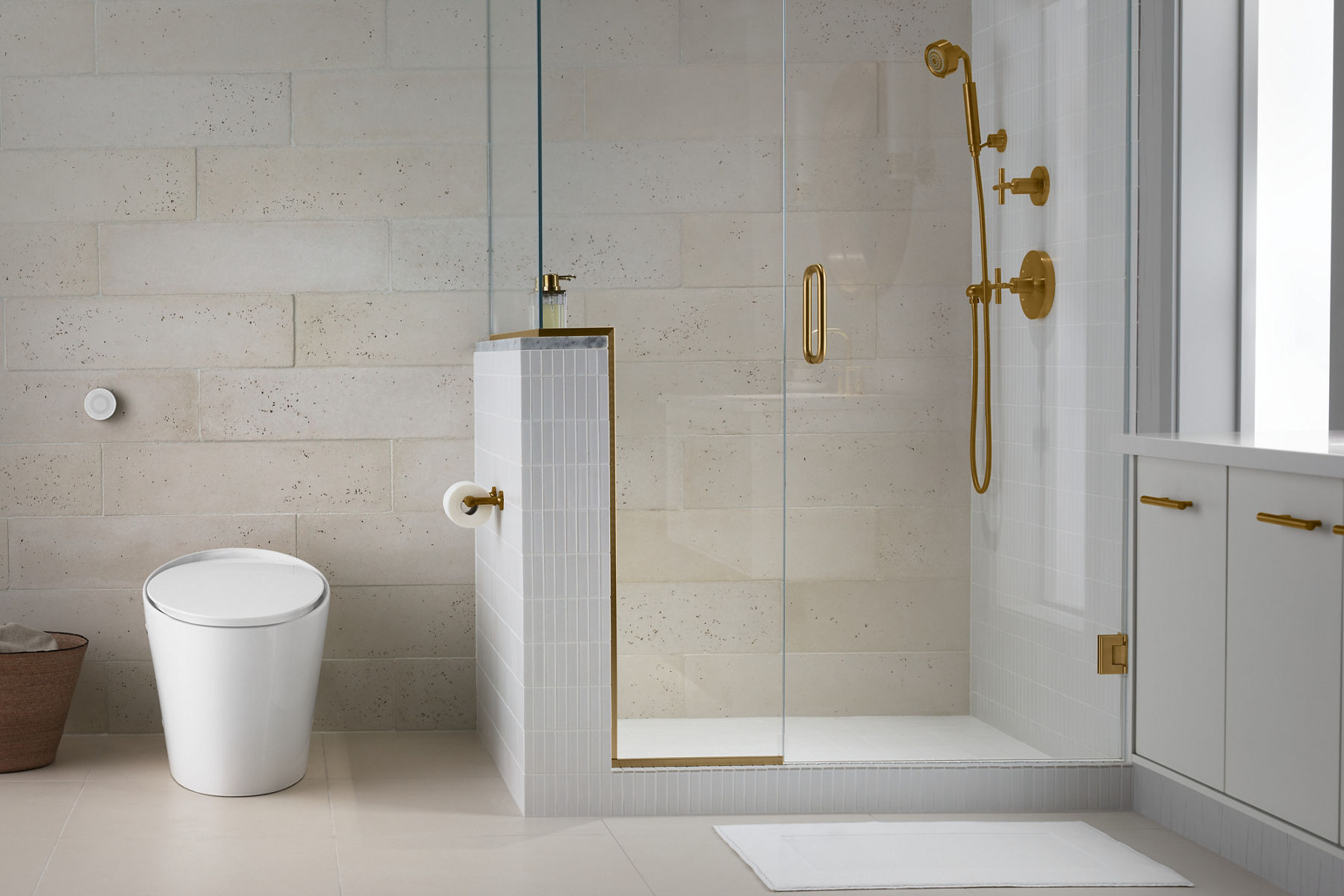 A white KOHLER elongated toilet with concealed tank in a bathroom with light tile walls and flor and large, glass-paneled walk-in shower.