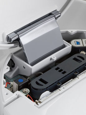 The interior electronic controls of a toilet's battery flush mechanism at the rear of the toilet under the lid.