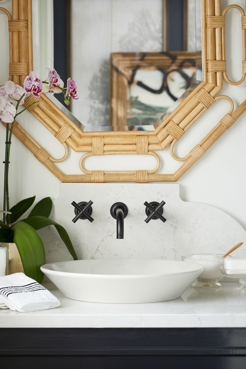 Une salle de bain avec un robinet déployé KOHLER mural avec poignées en croix, un lavabo en surface rond blanc, un dessus de meuble-lavabo blanc et un miroir octogonal à cadre en rotin.