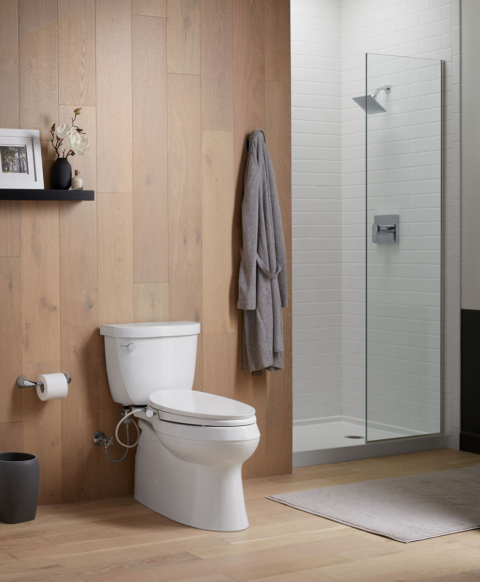 Une toilette allongée blanche KOHLER avec siège de bidet dans une salle de bain avec douche en alcôve et murs à panneaux en bois.