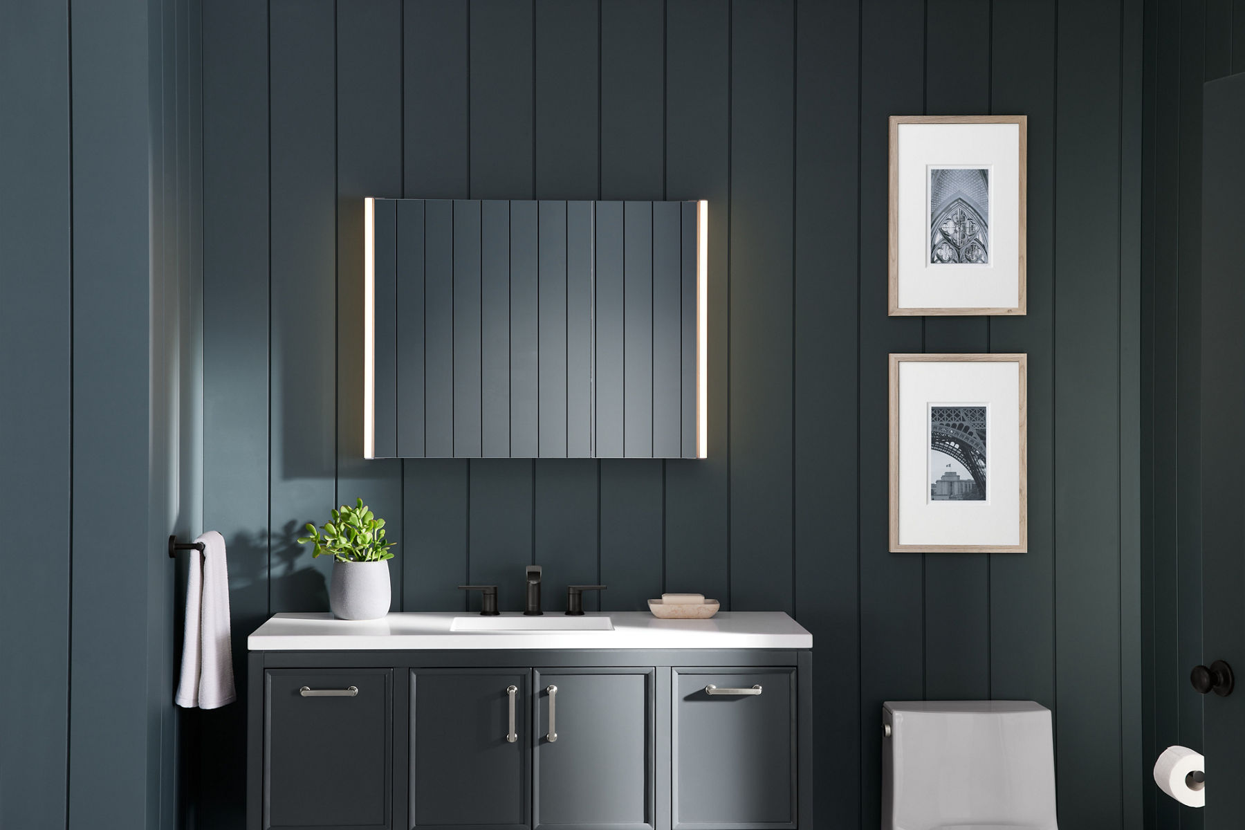 Une armoire à pharmacie éclairée KOHLER Verdera brille dans une salle de bain avec des murs gris foncé, un meuble-lavabo gris, un dessus de meuble-lavabo blanc, des toilettes blanches et des œuvres d’art encadrées avec des passe-partout blancs.